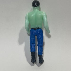 May include: A small plastic figurine of a person viewed from the back. The figure has a green torso, blue pants, black boots, and black hair. The figure is standing upright.