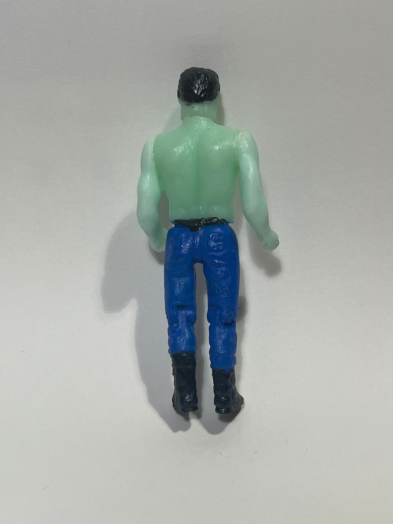 May include: A small plastic figurine of a person viewed from the back. The figure has a green torso, blue pants, black boots, and black hair. The figure is standing upright.
