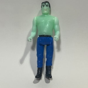 May include: A small plastic action figure of a muscular man with green skin, blue pants, and black boots. The figure has black hair and is posed with arms at its sides. The figure is standing on a white surface.