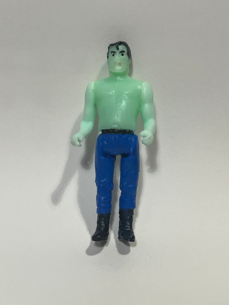 May include: A small plastic action figure of a muscular man with green skin, blue pants, and black boots. The figure has black hair and is posed with arms at its sides. The figure is standing on a white surface.