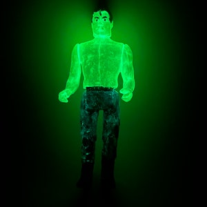 May include: A glowing green action figure of a man, with dark hair and a stern expression. The figure is wearing a light green shirt, blue pants, and black boots. The figure is illuminated against a dark background.