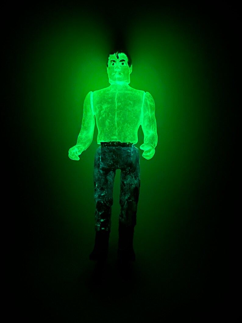 May include: A glowing green action figure of a man, with dark hair and a stern expression. The figure is wearing a light green shirt, blue pants, and black boots. The figure is illuminated against a dark background.