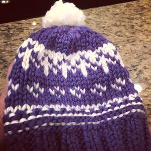 May include: Purple and white knitted hat with a white pom pom on top.