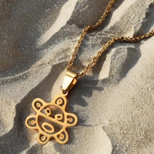 May include: Gold-colored necklace with a pendant featuring a stylized sun design. The pendant is suspended from a delicate chain. The necklace is displayed on a sandy surface, creating a contrast between the jewelry and the background.