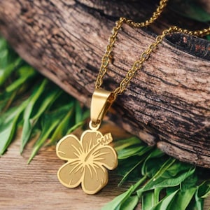 May include: A gold-colored necklace featuring a hibiscus flower pendant. The pendant is detailed with etched lines and hangs from a delicate chain. The necklace is displayed on a wooden surface with green leaves.