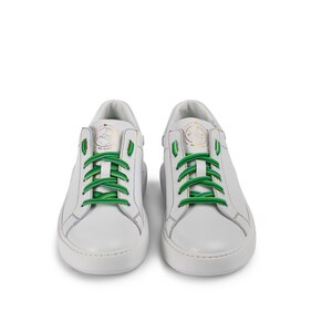 Puede incluir: Zapatillas bajas de cuero blanco con cordones verdes. Las zapatillas tienen una suela de goma blanca y un logotipo dorado en el lateral.