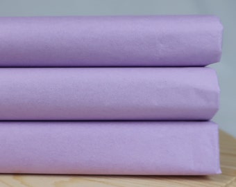 May include: Three rolls of lavender-colored paper are stacked on top of each other. The paper is a solid, light purple color. The rolls are resting on a light-colored wooden surface.