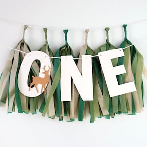 May include: White paper banner with the word "ONE" in large letters, decorated with green and brown tissue paper tassels. A small brown paper deer cutout is attached to the letter "O".