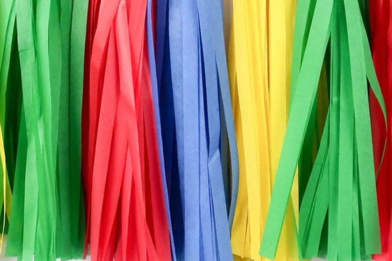 May include: A close-up of a colorful tissue paper streamer garland. The garland is made of green, red, blue, yellow, and green tissue paper streamers. The streamers are hanging down from a white background.