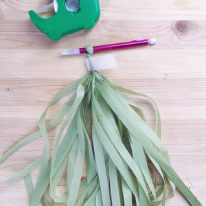 May include: A green tape dispenser with a roll of tape and a pink pencil next to a bundle of light green tissue paper streamers.