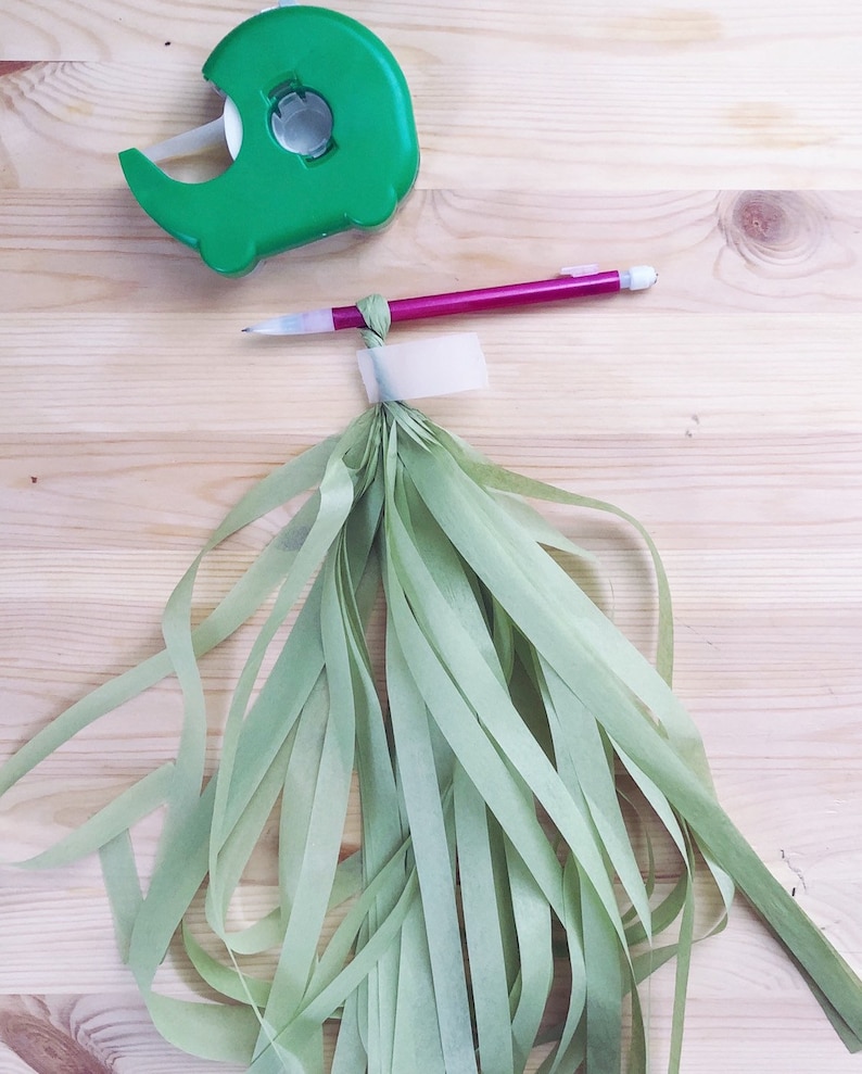 May include: A green tape dispenser with a roll of tape and a pink pencil next to a bundle of light green tissue paper streamers.