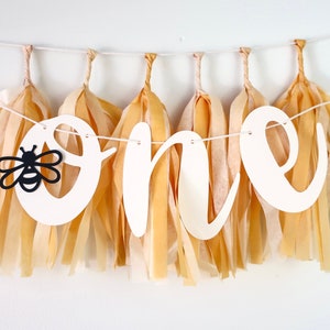 May include: A white banner with the word "one" in cursive script, decorated with black and yellow paper tassels and a black silhouette of a bee. The banner is perfect for a first birthday party.
