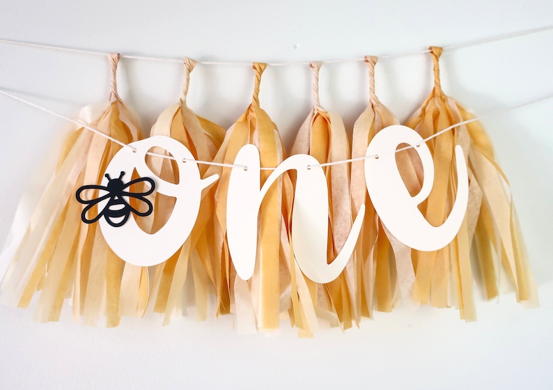 May include: A white banner with the word "one" in cursive script, decorated with black and yellow paper tassels and a black silhouette of a bee. The banner is perfect for a first birthday party.