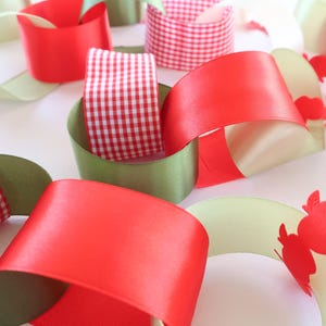 Op de afbeelding: Een decoratieve papieren kettingslinger met in elkaar grijpende lussen van rood satijnlint, rood-wit gingham lint en olijfgroen papier. Sommige delen bevatten een uitgestanst patroon van kleine rode appeltjes langs de randen van de lussen.