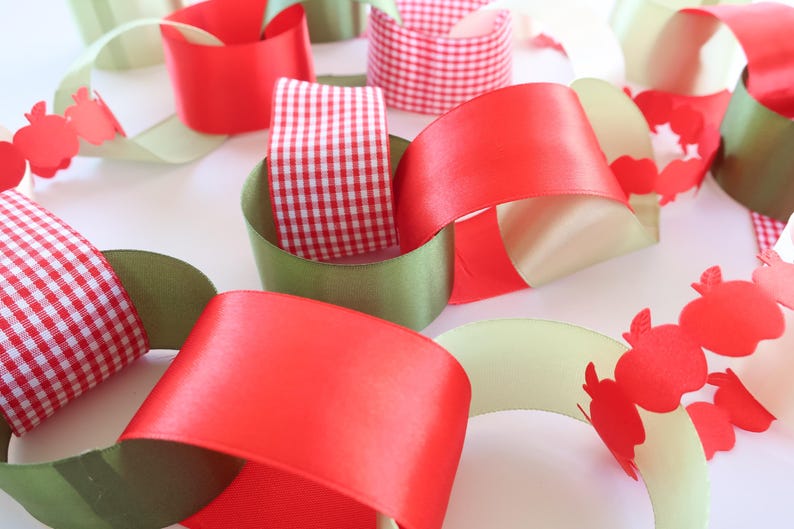 Op de afbeelding: Een decoratieve papieren kettingslinger met in elkaar grijpende lussen van rood satijnlint, rood-wit gingham lint en olijfgroen papier. Sommige delen bevatten een uitgestanst patroon van kleine rode appeltjes langs de randen van de lussen.