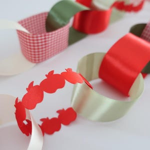 Op de afbeelding: Een decoratieve papieren slinger met in elkaar grijpende lussen in rood, wit en olijfgroen. De slinger bevat segmenten van rood-wit geblokt papier, een effen rood satijnachtig lint en een rode papieren strook uitgesneden in de vorm van appels.
