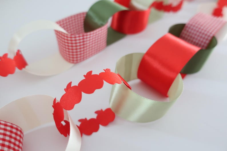 Op de afbeelding: Een decoratieve papieren slinger met in elkaar grijpende lussen in rood, wit en olijfgroen. De slinger bevat segmenten van rood-wit geblokt papier, een effen rood satijnachtig lint en een rode papieren strook uitgesneden in de vorm van appels.