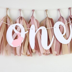 May include: A white banner with the word "one" in cursive script, decorated with pink and beige tissue paper tassels. A pink bunny cutout is attached to the "o".