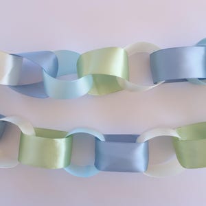 May include: Two decorative paper chains in pastel colors. The chains are made of interlocking loops of satin ribbon in shades of light blue, green, and cream. The chains are arranged horizontally against a white background.