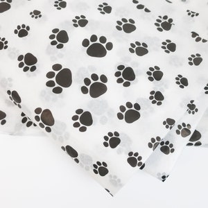 May include: White tissue paper with a repeating pattern of black paw prints. The paw prints vary in size and are scattered across the paper. The tissue paper is folded, showing multiple layers.