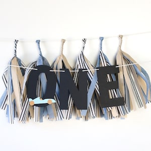 May include: A black and white banner with the word "ONE" in large black letters. The banner is decorated with blue and tan striped paper tassels. A small blue race car is attached to the letter "O".