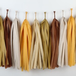 May include: A garland of tissue paper tassels in shades of brown, beige, and yellow. The tassels are strung together with a white string and hang down in a row.