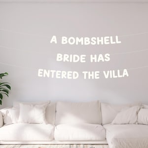 May include: A white banner with the text "A BOMBSHELL BRIDE HAS ENTERED THE VILLA" hangs above a white sectional sofa. A potted plant sits to the left. The room is painted white.