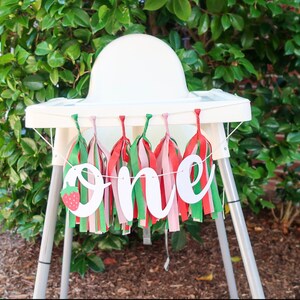 May include: A white high chair with a banner that says "one" in white script letters with a red strawberry graphic. The banner is decorated with green, red, and pink tassels.