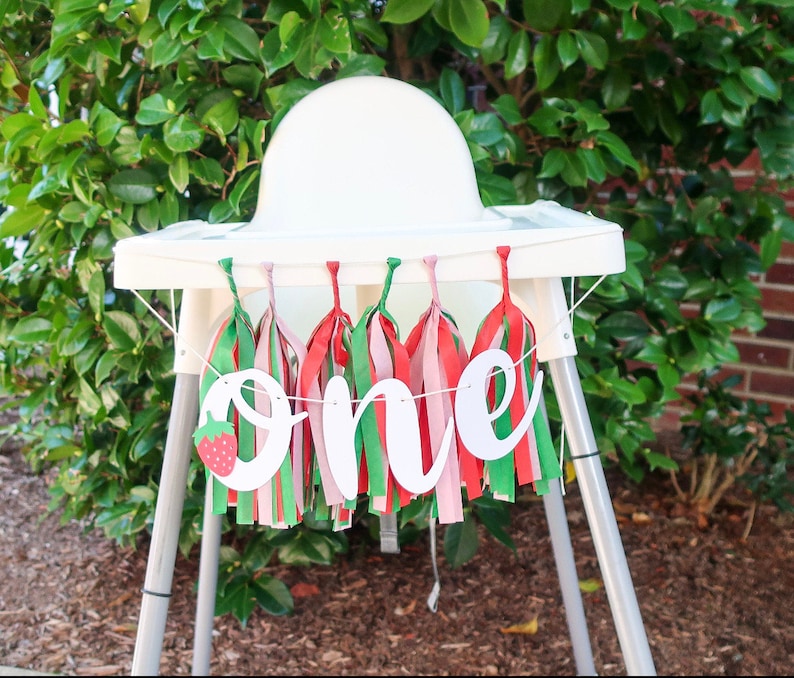 May include: A white high chair with a banner that says "one" in white script letters with a red strawberry graphic. The banner is decorated with green, red, and pink tassels.