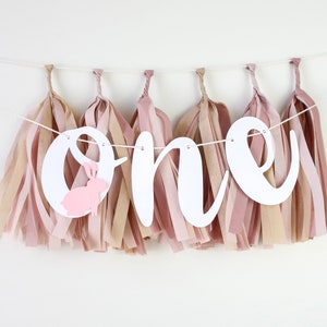 May include: White banner with the word "one" in cursive, decorated with pink and beige tissue paper tassels. A small pink bunny cutout is attached to the "o".