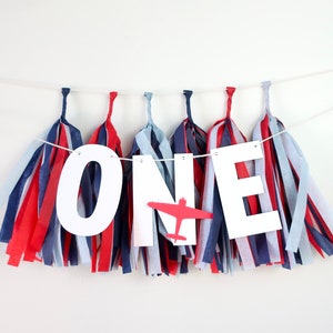 May include: A celebratory banner for a first birthday. The banner spells "ONE" in white letters, with a red airplane graphic. The banner is decorated with red, white, and blue tissue paper tassels, creating a patriotic theme.