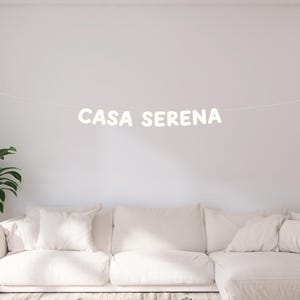 May include: White banner with the text "CASA SERENA" hanging on a white wall above a white couch with white pillows.