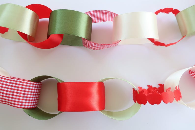 Op de afbeelding: Een decoratieve papieren kettingslinger met in elkaar grijpende lussen van diverse materialen. De schakels bestaan uit effen rode, olijfgroene en cr&egrave;mekleurige satijnen linten, rood-wit geblokt papier en rode papieren uitsparingen in de vorm van appels.