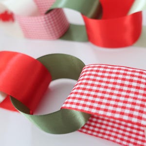 Op de afbeelding: Een decoratieve ketting gemaakt van in elkaar grijpende lussen van lint. De ketting heeft een herhalend patroon van effen rode satijn, effen olijfgroene satijn, witte stroken en rood-wit geblokte vichy-stof die met elkaar verbonden zijn.