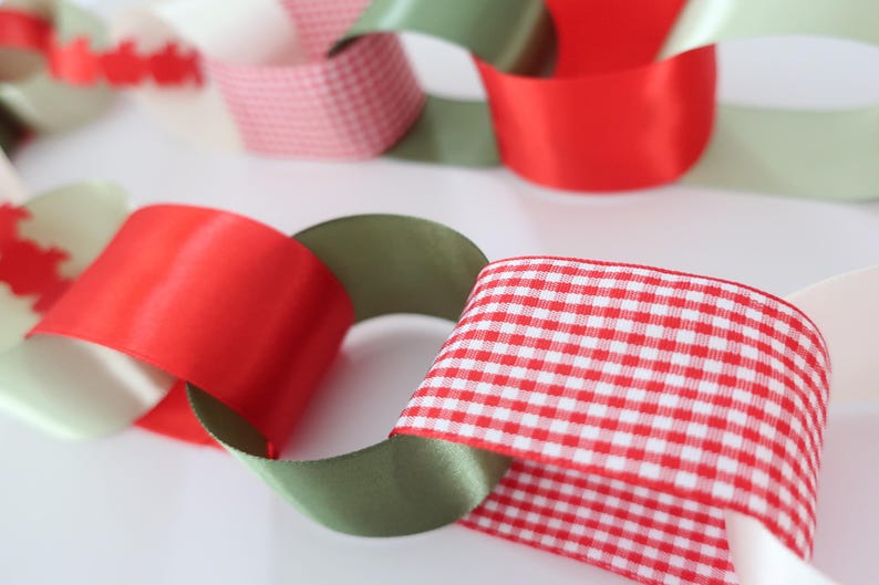 Op de afbeelding: Een decoratieve ketting gemaakt van in elkaar grijpende lussen van lint. De ketting heeft een herhalend patroon van effen rode satijn, effen olijfgroene satijn, witte stroken en rood-wit geblokte vichy-stof die met elkaar verbonden zijn.