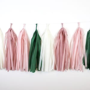 May include: A garland of tissue paper tassels in shades of blush pink, ivory white, and forest green. The tassels are strung together on a white string.