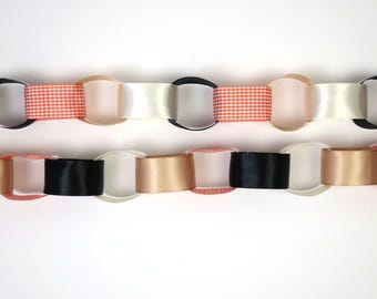 Könnte beinhalten: Zwei horizontale Papierketten aus ineinandergreifenden Schlaufen. Die obere Kette besteht aus abwechselnden schwarzen, weißen und orangefarbenen, karierten Papierschlaufen. Die untere Kette besteht aus abwechselnden schwarzen, beigen und orangefarbenen Schlaufen.