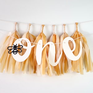 May include: A white paper banner with the word "one" in cursive letters, decorated with tissue paper tassels in a light beige color. A black silhouette of a bee is on the first letter.