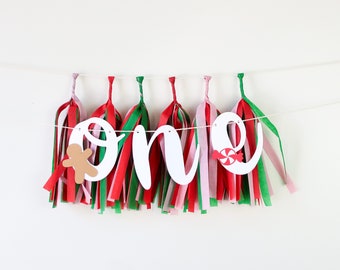 Puede incluir: Pancarta de papel blanco con la palabra "one" en letras cursivas. La pancarta está decorada con borlas de papel de seda rojo, verde y rosa. Hay un hombre de jengibre y un bastón de caramelo en la pancarta.