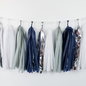 May include: A garland of tissue paper tassels in navy blue, silver, and light gray. The tassels are strung together with white string.