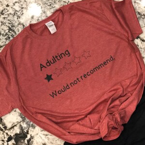 以下が含まれることがあります： 「Adulting Would not recommend」のテキストが入った赤いTシャツ。デザインには黒い星と4つのアウトラインの星が含まれており、評価システムを示唆しています。シャツは柔らかい素材で作られています。