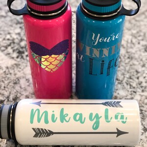 May include: Three insulated water bottles. One is pink with a mermaid heart design, another is blue with the text "You're winning at life", and the third is white with the name "Mikayla" and arrow designs. All have black lids.