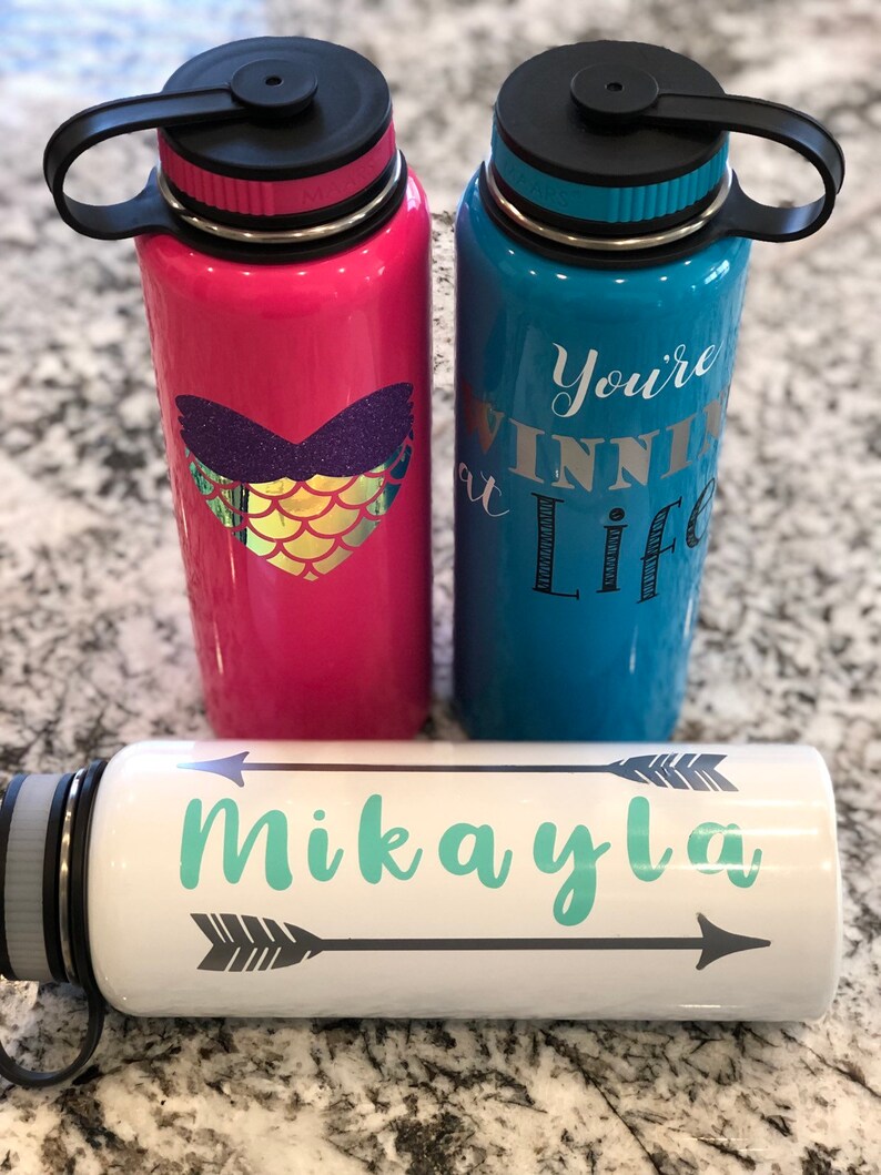 May include: Three insulated water bottles. One is pink with a mermaid heart design, another is blue with the text "You're winning at life", and the third is white with the name "Mikayla" and arrow designs. All have black lids.
