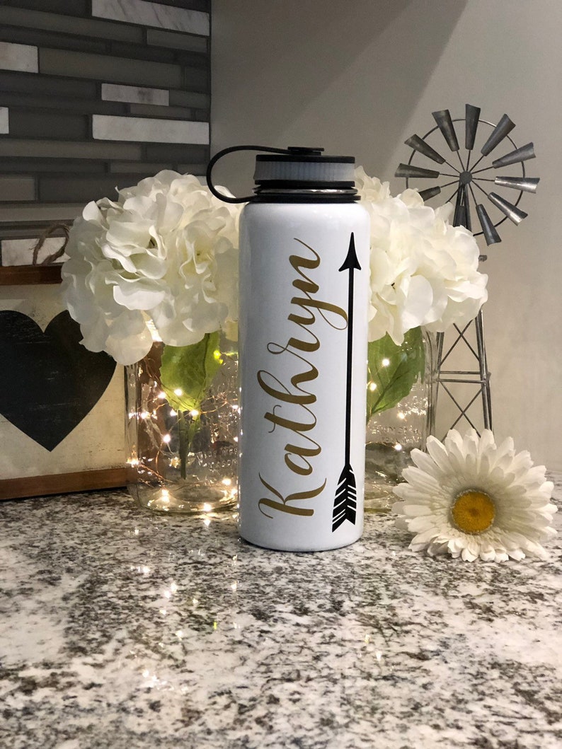 May include: White insulated water bottle with a black lid and handle. The bottle features the name "Kathryn" in gold script with a black arrow design. The bottle is surrounded by white flowers and decorative elements.