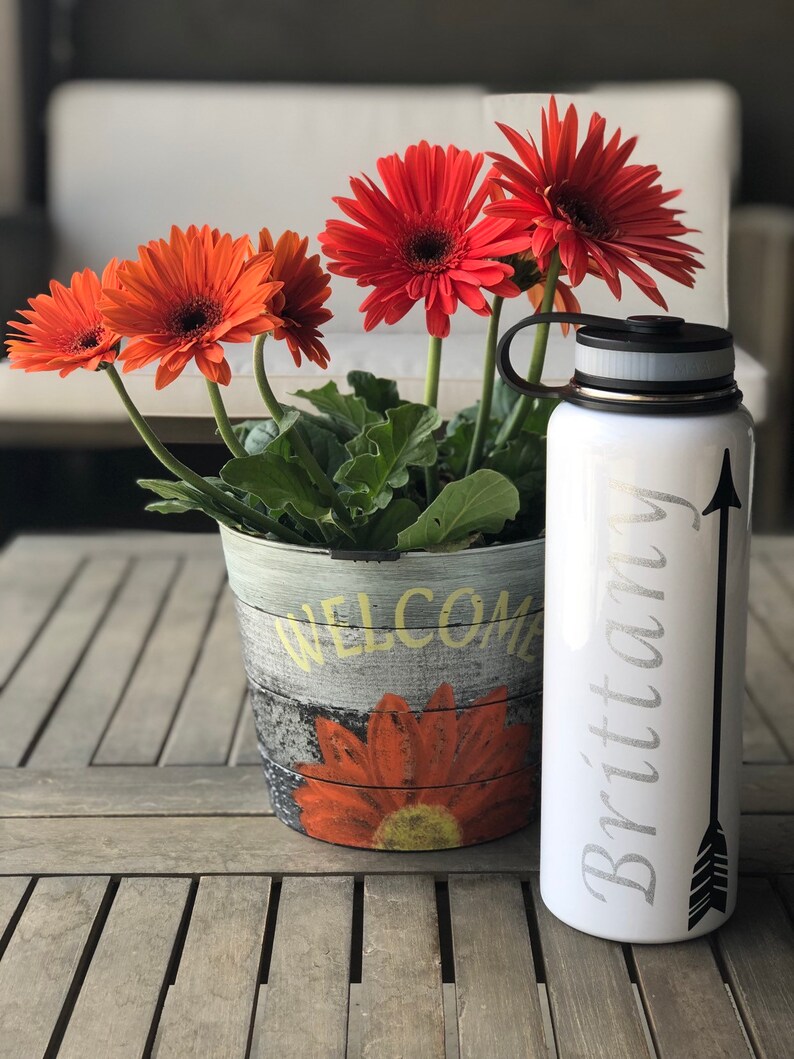 May include: A white water bottle with the name "Brittany" and an arrow design. Beside it is a flower pot with orange gerbera daisies and the word "WELCOME". The pot has a painted sunflower design.