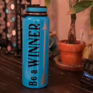 May include: A turquoise water bottle with a black lid and the words "Be a WINNER" in black, with a silver arrow design. The bottle is cylindrical and appears to be made of metal.