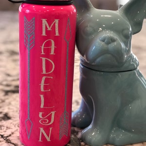 May include: A pink water bottle with the name "MADELYN" in silver glitter lettering, flanked by teal arrow designs. The bottle has a black lid and a black handle. A gray ceramic bulldog figurine is next to the bottle.
