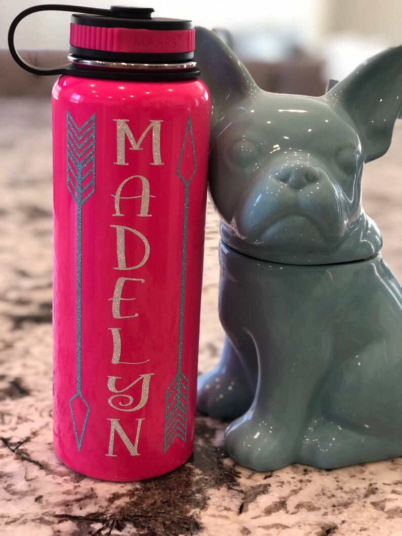 May include: A pink water bottle with the name "MADELYN" in silver glitter lettering, flanked by teal arrow designs. The bottle has a black lid and a black handle. A gray ceramic bulldog figurine is next to the bottle.