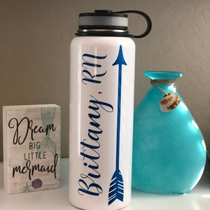 May include: White water bottle with a black lid and handle, featuring a blue arrow design and the name "Brittany, RN" in a script font. A small wooden block with mermaid-themed text and a blue vase are also visible.