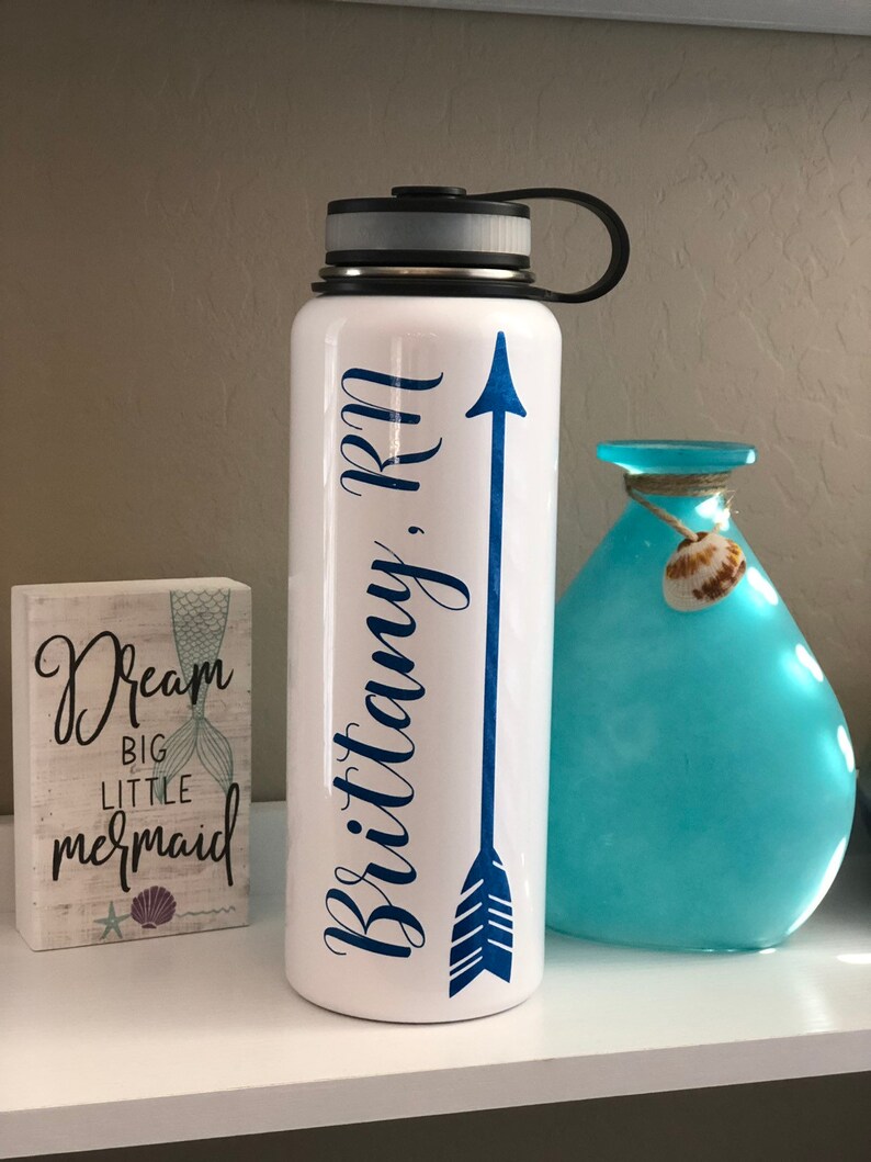 May include: White water bottle with a black lid and handle, featuring a blue arrow design and the name "Brittany, RN" in a script font. A small wooden block with mermaid-themed text and a blue vase are also visible.